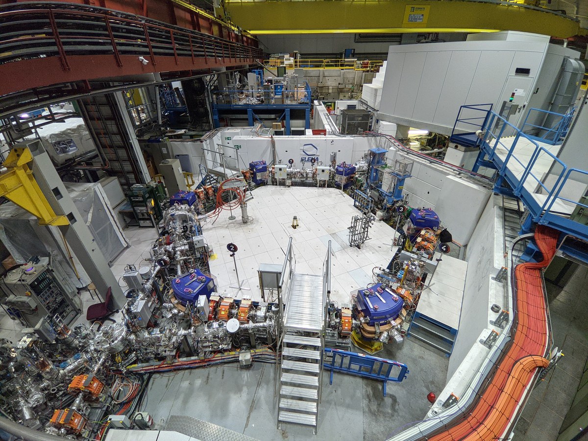 Vue plongeante sur le hall du décélérateur ELENA au CERN : un anneau d'aimants bleus et orange posé au sol, entouré de passerelles métalliques et de câbles
