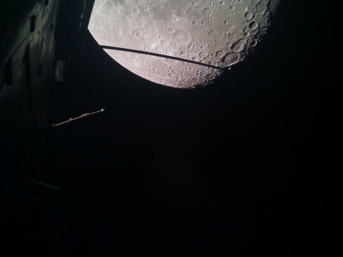 Surface grise de la Lune criblée de cratères, vue en partie depuis la sonde Juice, avec une antenne et un mât de la sonde au premier plan dans le noir