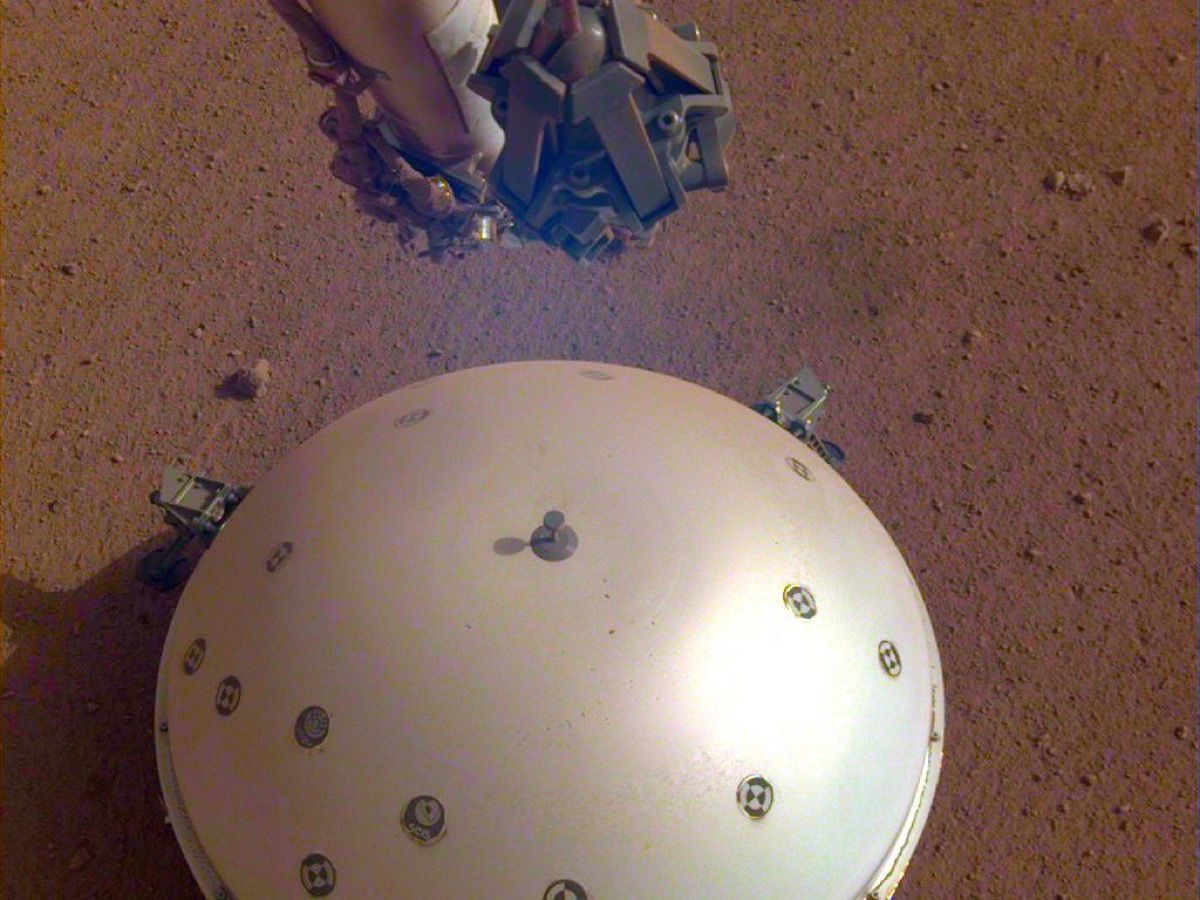 Coupole blanche posée sur le sol rouge de Mars, entourée de petits cailloux, avec la pince du bras robotique juste au-dessus