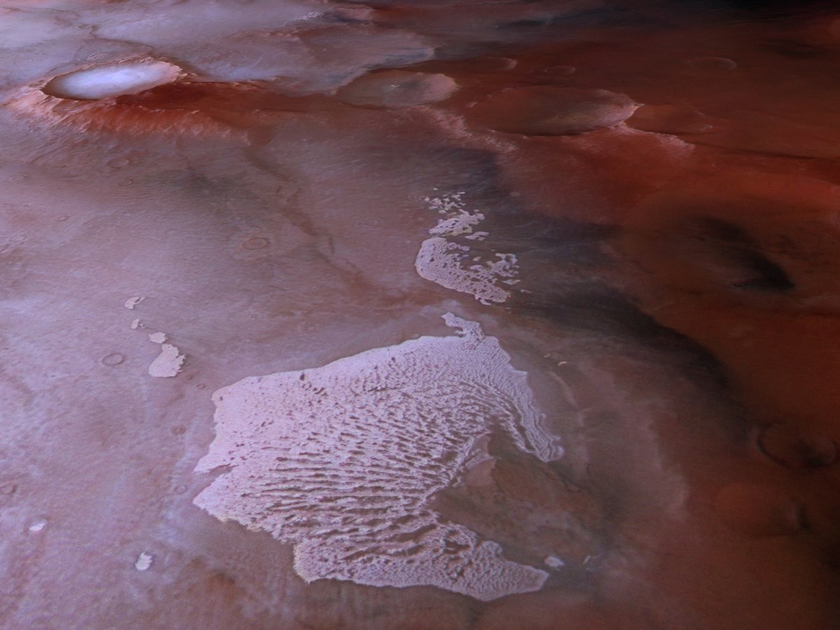 Vue en perspective d'une plaine martienne rosée et rouge sombre, avec des cratères givrés et une grande plaque de dunes blanches parcourue de rides