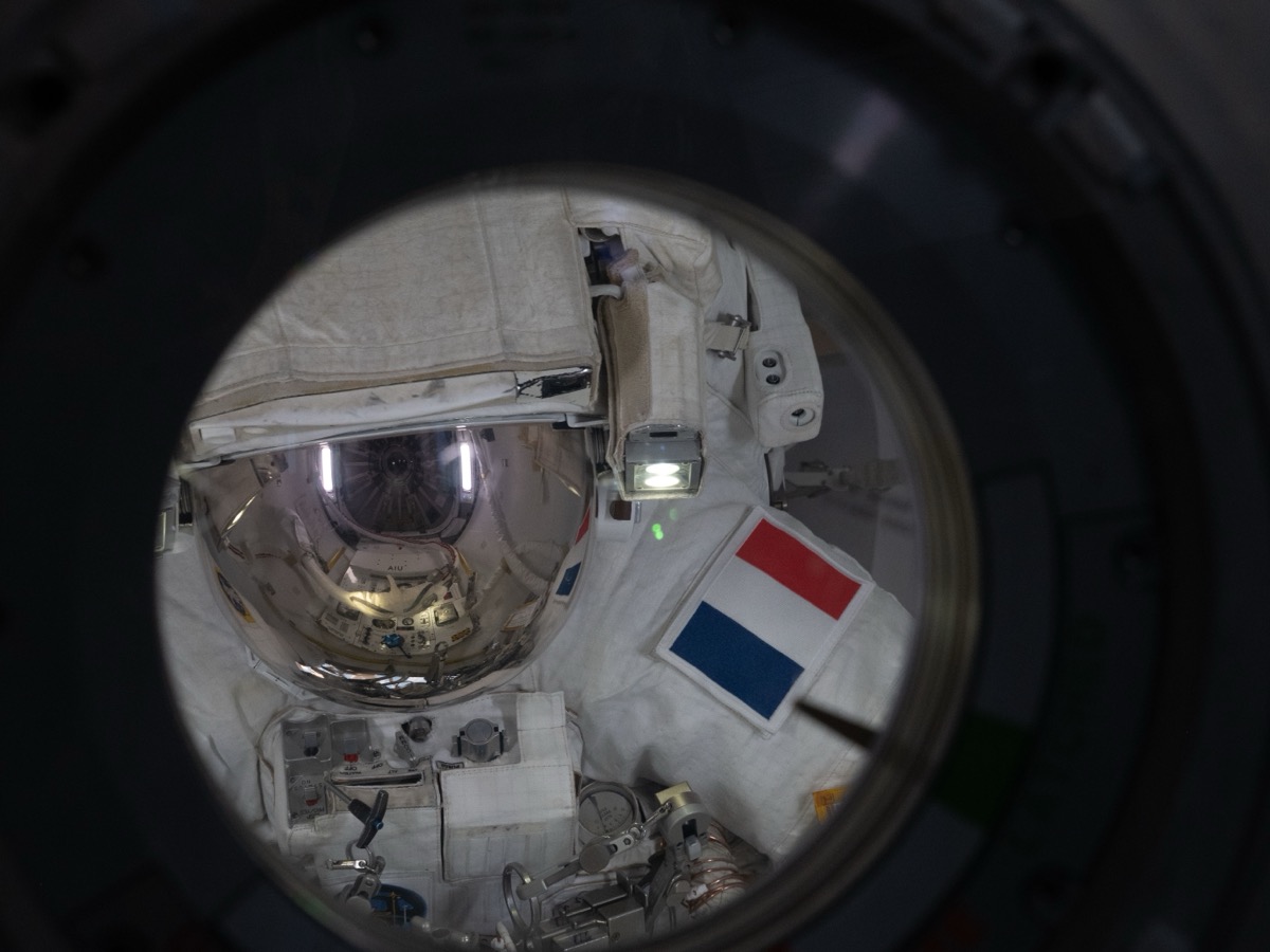 Astronaute en scaphandre blanc, visière dorée baissée et écusson tricolore sur la manche, vue à travers le hublot rond d’un sas