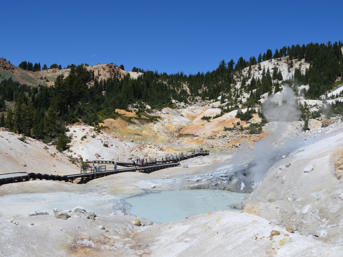 Vue d'ensemble d'un bassin volcanique blanc et ocre avec un plan d'eau laitée, des panaches de vapeur et une passerelle en bois qui traverse le site
