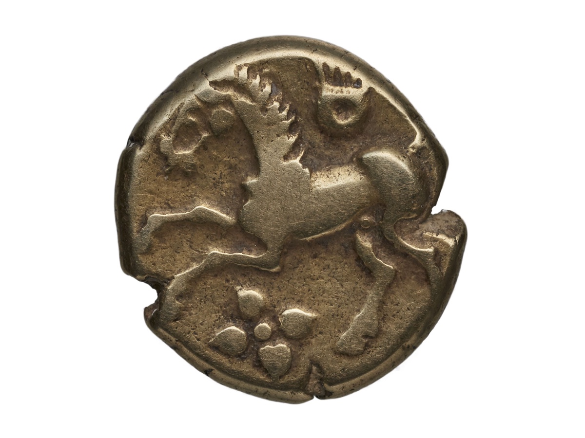 Pièce gauloise en or pâle, de forme irrégulière, frappée d'un cheval stylisé au galop au-dessus d'un motif en forme de fleur, sur fond blanc