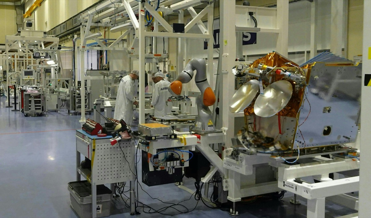 Satellite OneWeb en cours d'assemblage sur un bâti blanc dans une vaste salle d'intégration, avec un bras robotisé gris et orange au premier plan et deux techniciens en blouse et charlotte qui travaillent sur des postes en arrière-plan