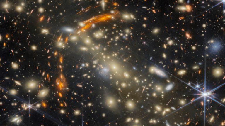 Amas de galaxies dorées sur fond noir, entouré d'arcs orangés étirés par la lentille gravitationnelle