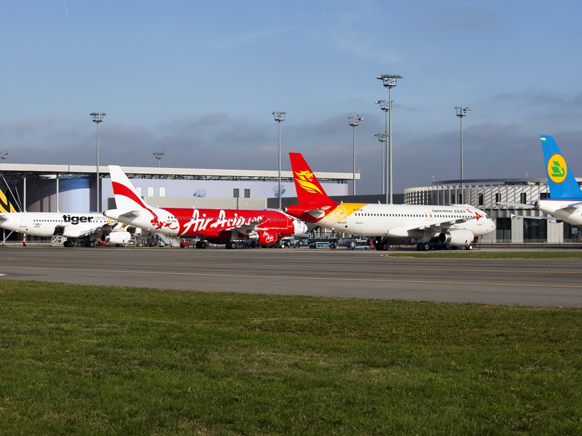 Plusieurs Airbus A320 aux couleurs de compagnies asiatiques alignés sur le tarmac devant un bâtiment vitré de l’aéroport de Toulouse-Blagnac, pelouse au premier plan