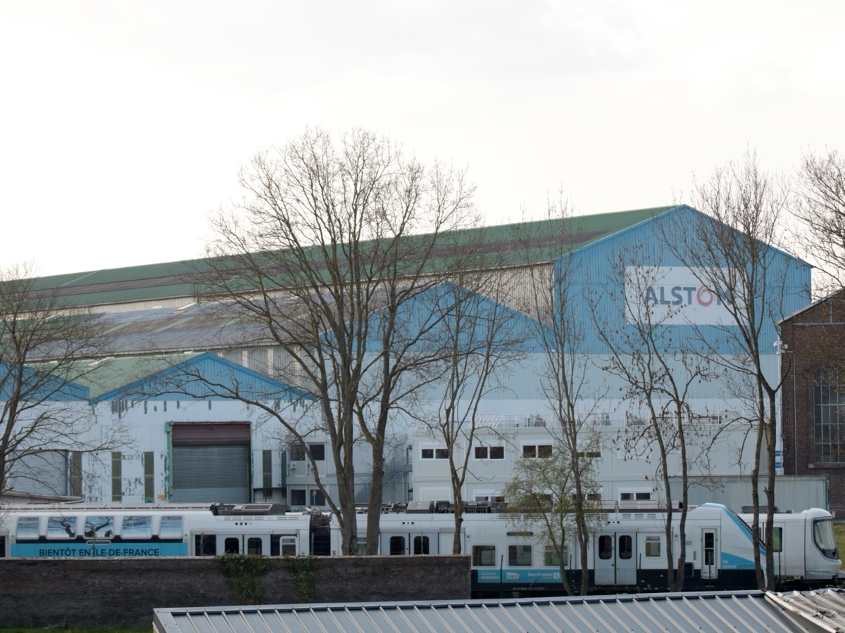 Grands bâtiments industriels bleus et blancs du site Alstom de Petite-Forêt, avec deux rames de train neuves garées devant, derrière une rangée d’arbres sans feuilles
