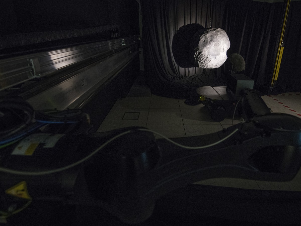Salle d’essai assombrie avec un rail métallique, un bras robotisé et une maquette éclairée par une source unique