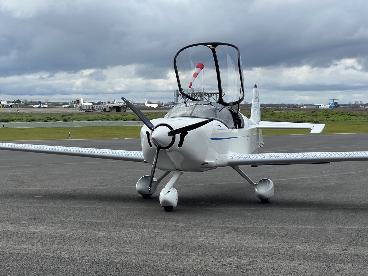 Petit avion blanc à aile basse, hélice bipale et verrière relevée, stationné de face sur le tarmac d’un aéroport sous un ciel gris