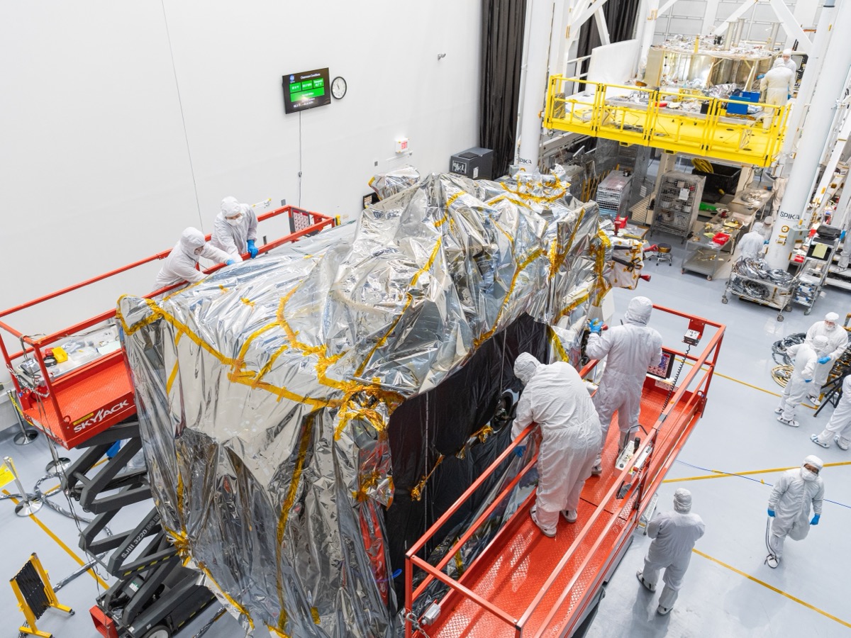 Techniciens en combinaison blanche autour d'un satellite emballé dans une couverture isolante dorée et argentée, dans une salle blanche