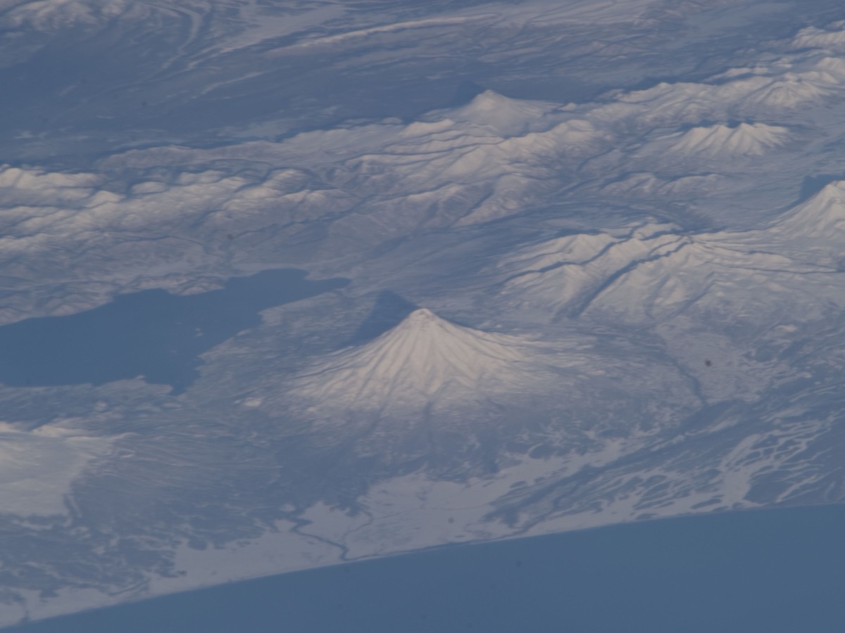 Volcans enneigés d’une péninsule côtière vus de haut, avec un cône régulier au centre et la mer sombre à gauche