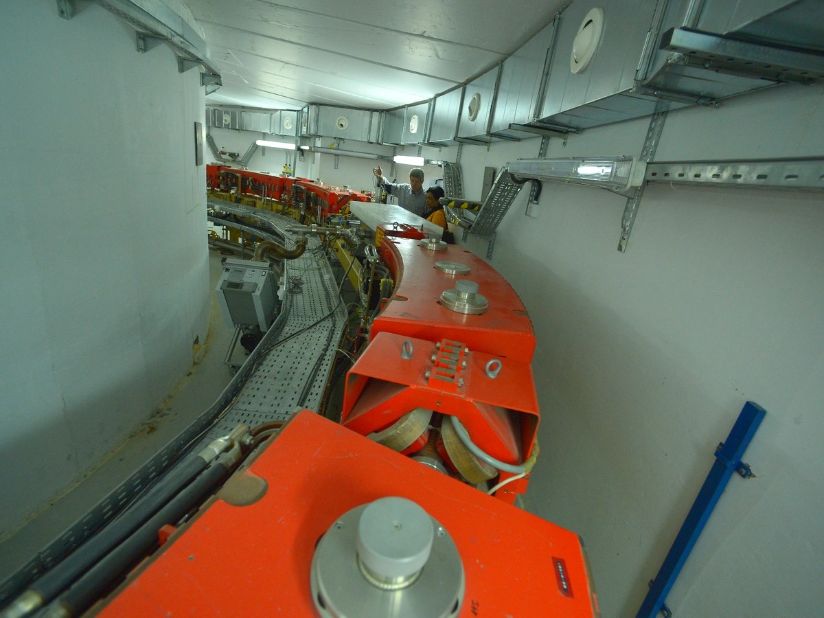 Série d’électroaimants orange disposés en courbe dans l’anneau intérieur d’un synchrotron, le long d’un couloir de béton blanc, avec deux techniciens au travail à l’arrière-plan
