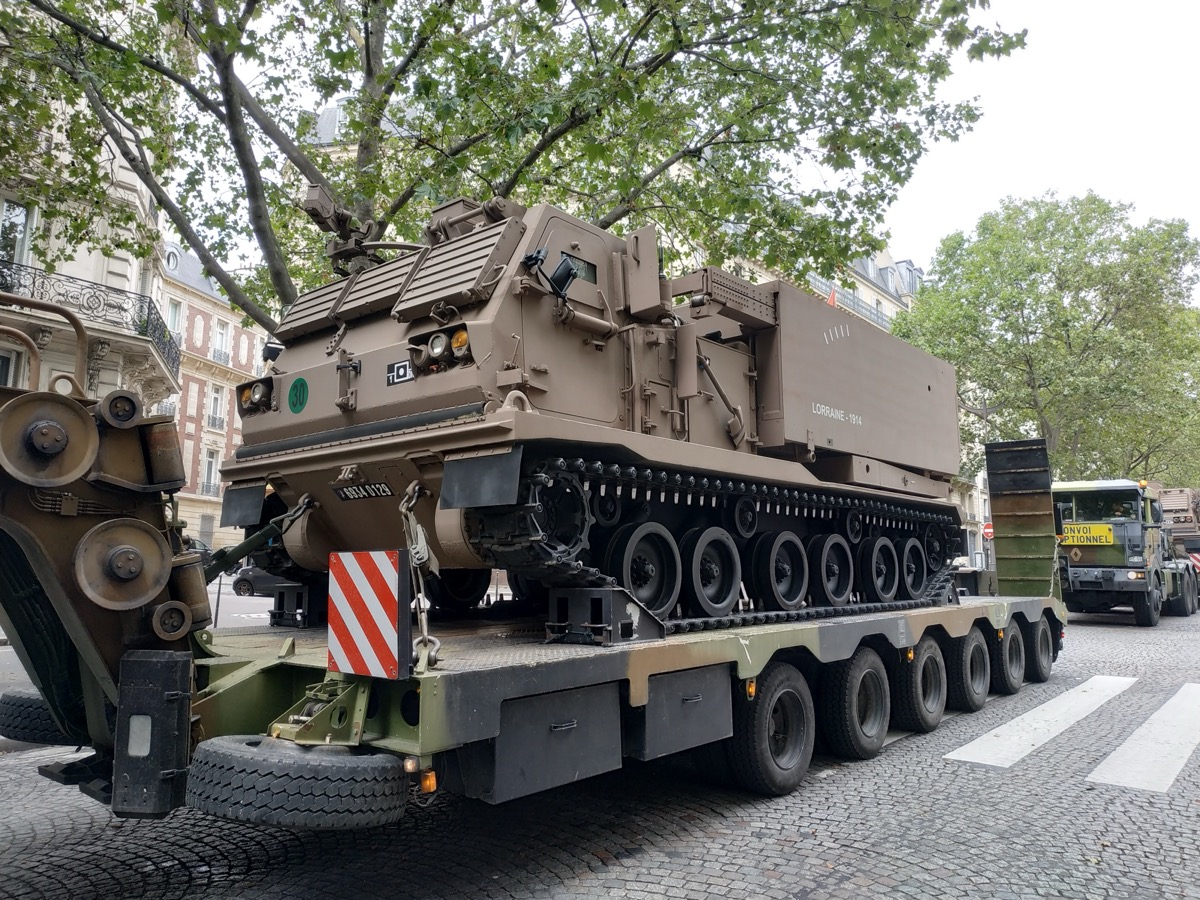 Lance-roquettes unitaire de l'armée de terre, engin chenillé de couleur sable, sanglé sur une remorque porte-chars tractée dans une rue bordée d'immeubles haussmanniens