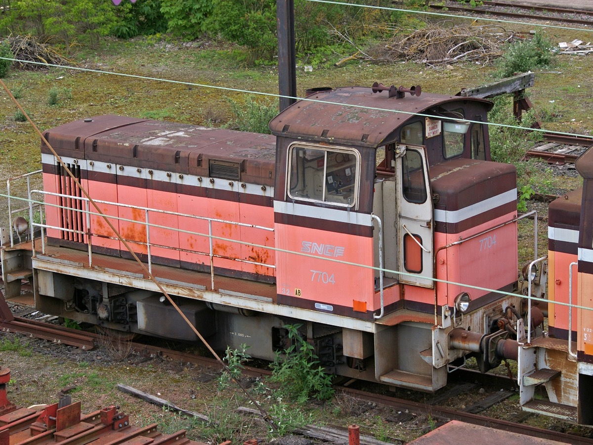 Locomotive de manoeuvre diesel SNCF Y 7704 rouge et blanche, carrosserie rouillée, garée sur une voie envahie d'herbe dans un faisceau de triage