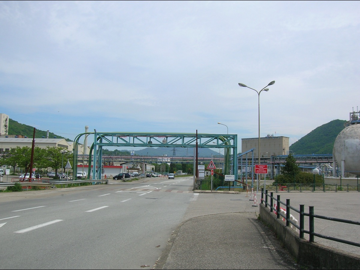 Passerelle de tuyauteries vert-de-gris enjambant la route à l'entrée de la plateforme chimique de Basse-Jarrie, sphère de stockage blanche à droite