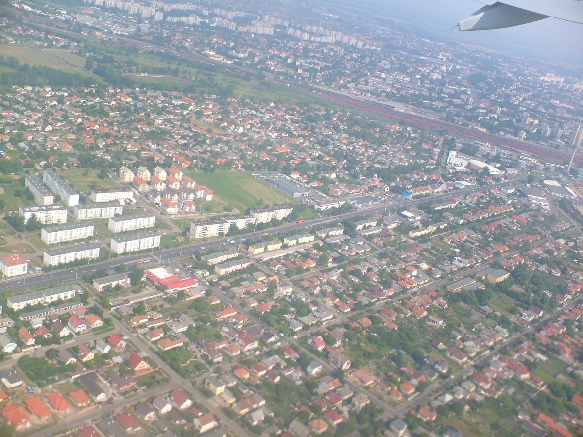 Vue aérienne d’une ville de plaine : barres d’immeubles, maisons aux toits rouges, voies ferrées et champs, avec l’aile de l’avion en haut à droite