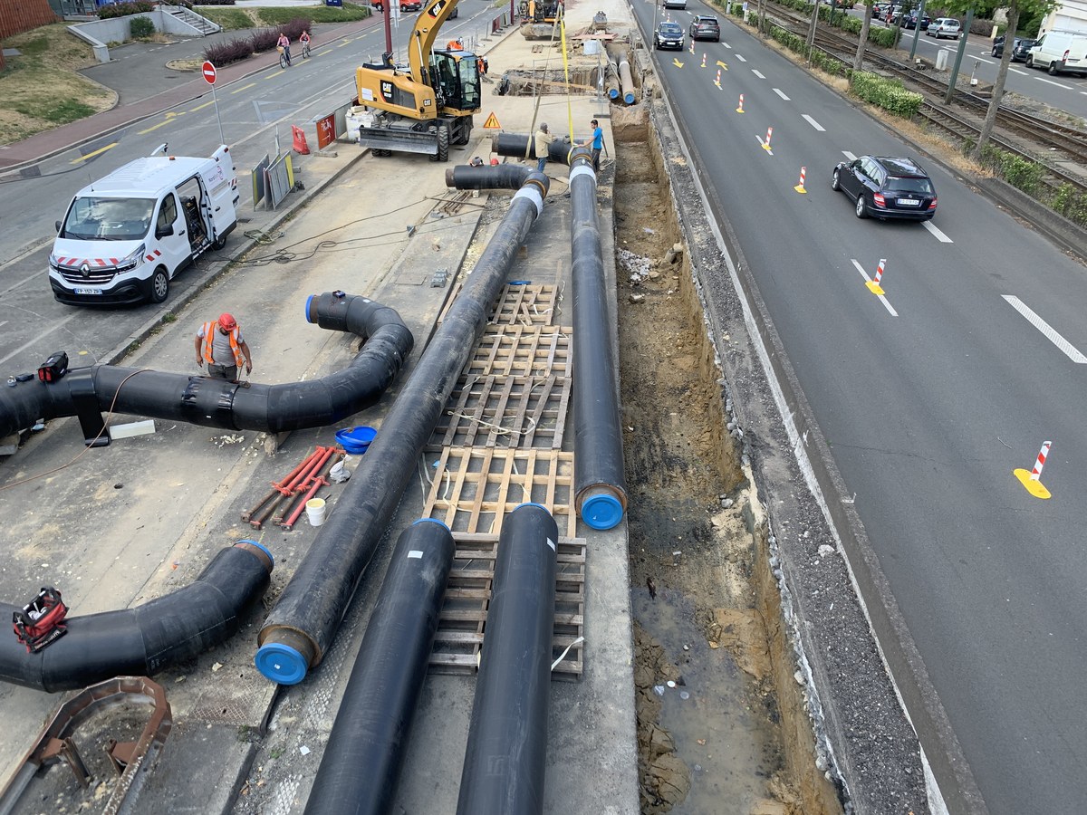 Tranchée de chantier le long d’une route à plusieurs voies, avec de gros tubes noirs aux embouts bleus posés sur des palettes, une pelleteuse jaune et deux ouvriers au travail