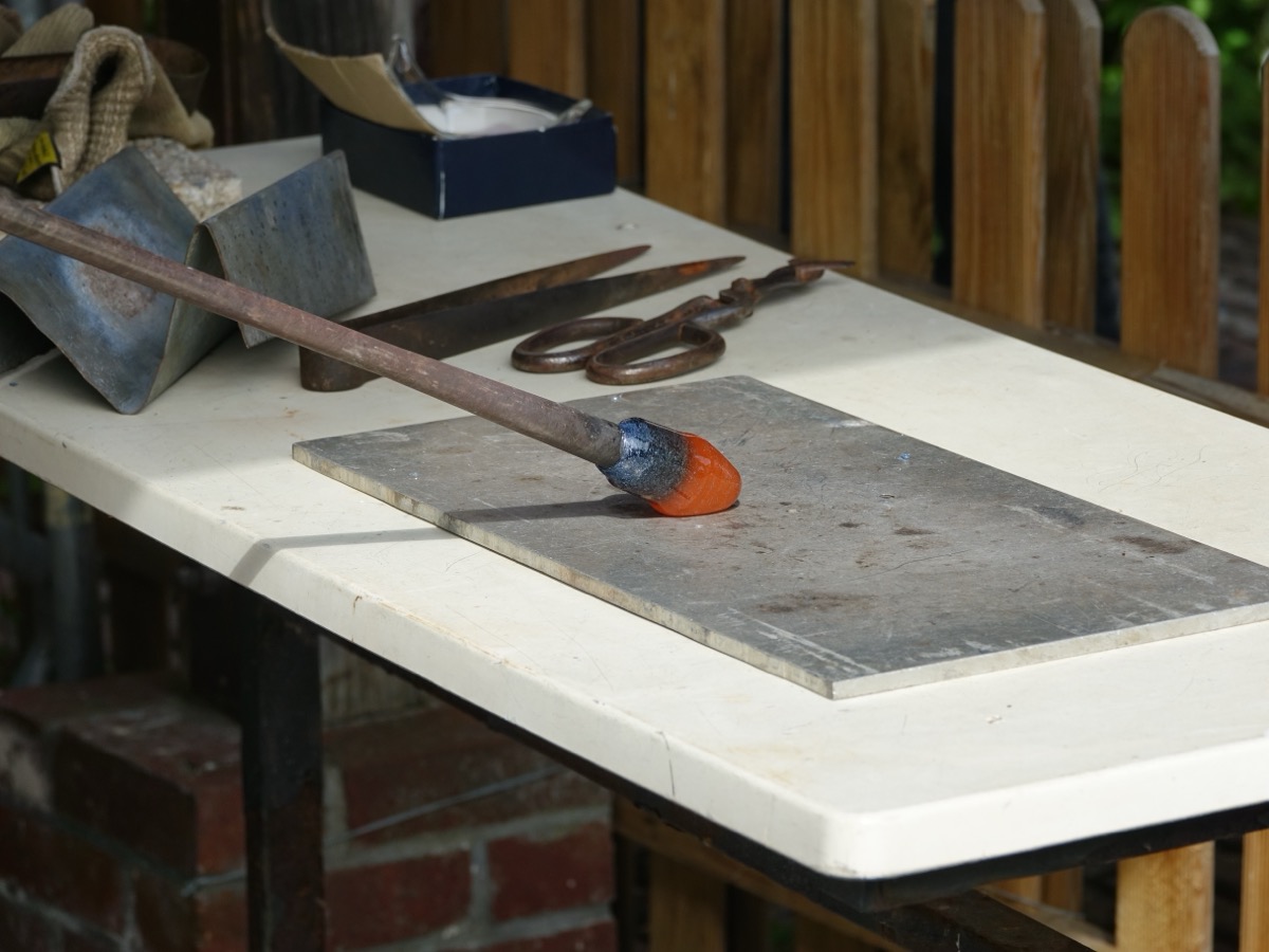 Canne de verrier posée sur une plaque de marbre, avec une masse de verre en fusion orange à son extrémité, à côté de ciseaux et de mailloches en bois sur un établi blanc