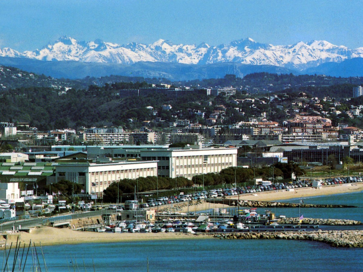 Bâtiments blancs du Centre spatial de Cannes Mandelieu en bord de mer, derrière un petit port de plaisance, avec des collines boisées et les Alpes enneigées à l’horizon