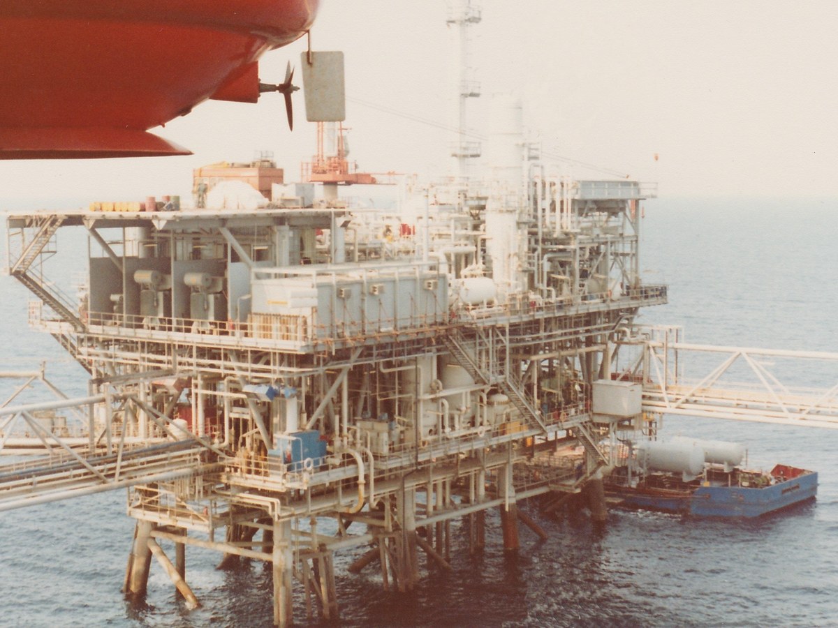 Plateforme pétrolière offshore blanche aux nombreuses tuyauteries, reliée par une passerelle, avec une barge amarrée à ses pieds sur une mer calme