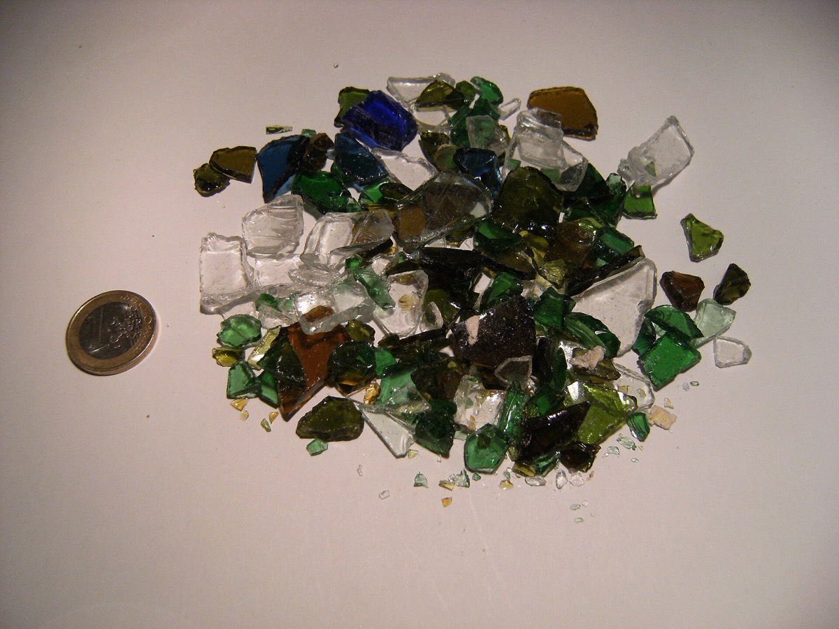 Poignée de morceaux de verre brisé vert, brun et incolore posés sur une surface claire, à côté d'une pièce de un euro