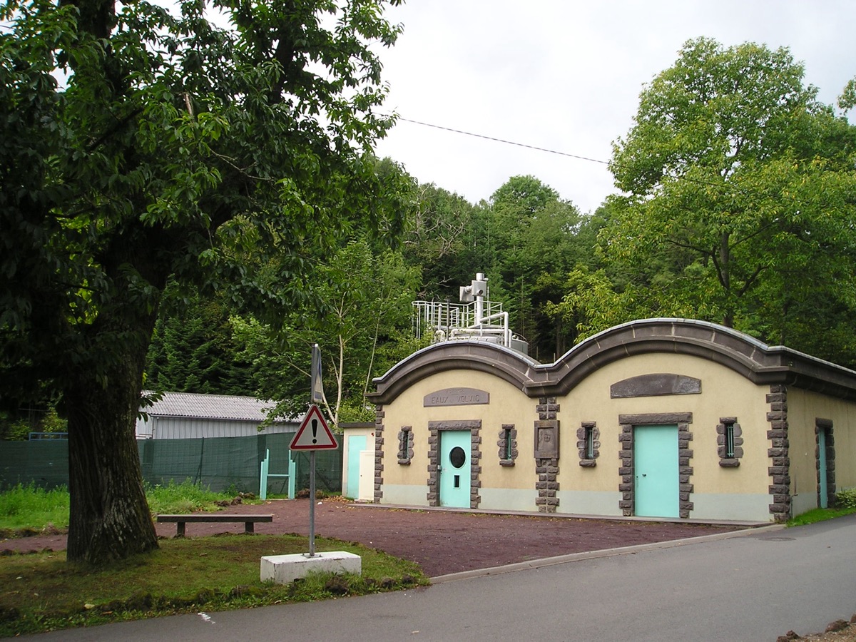 Bâtiment de captage des eaux de Volvic en pierre volcanique, deux portes vertes sous un toit ondulé, entouré d'arbres