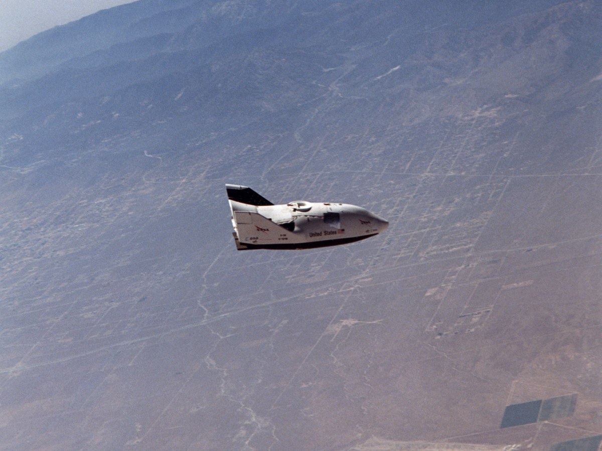 Petit engin spatial blanc à fuselage porteur et dérive noire, en vol plané au-dessus d’un désert quadrillé de routes, montagnes à l’horizon