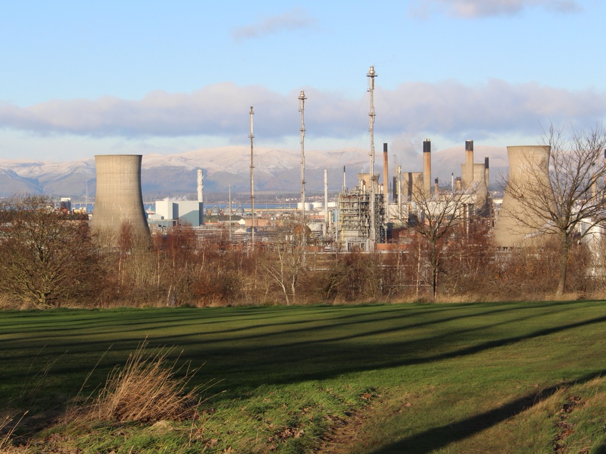 Vue lointaine d'un site pétrochimique avec tours de refroidissement et cheminées, devant des collines enneigées et une prairie au premier plan