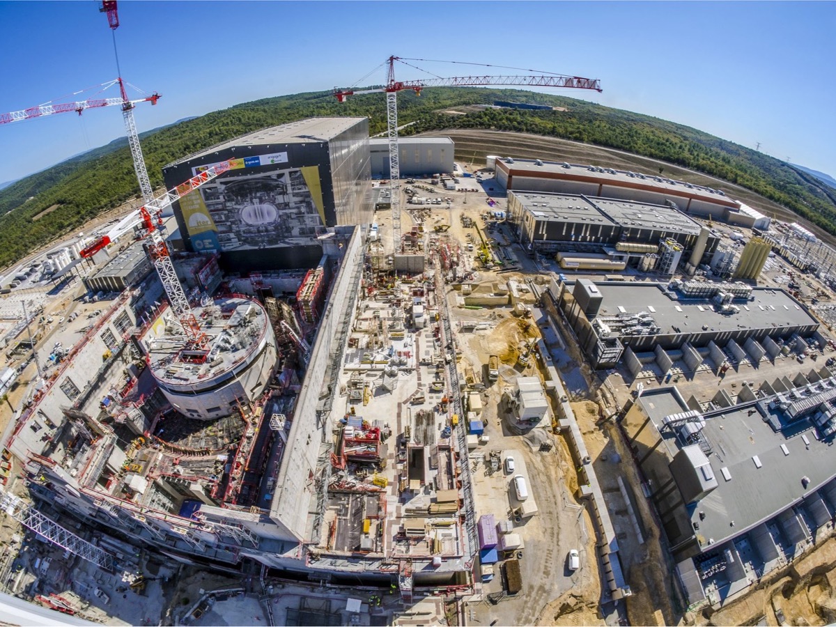 Vue aérienne du chantier ITER en Provence, avec les grues, le bâtiment tokamak en construction et les halls d'assemblage entourés de collines boisées