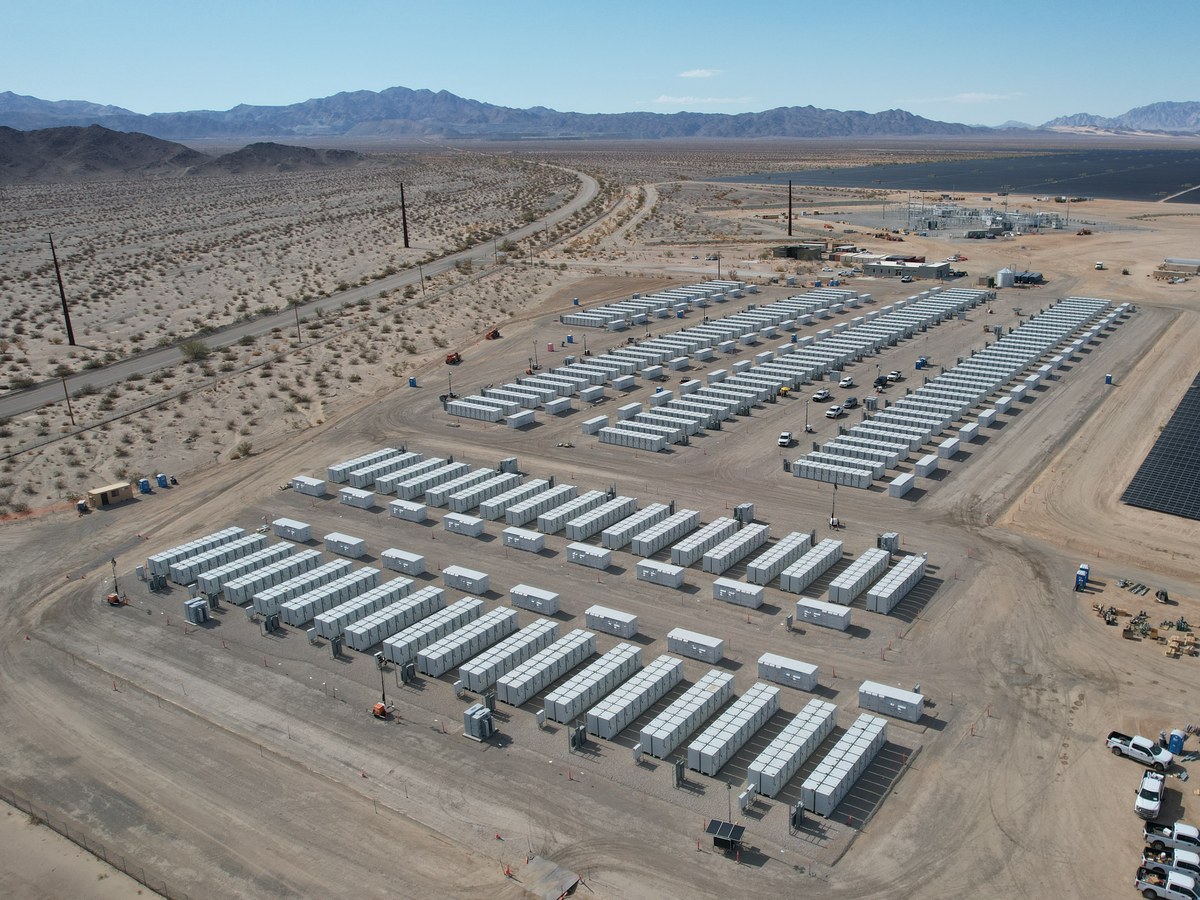 Vue aérienne de dizaines de conteneurs de batteries blancs alignés en rangées dans le désert, à côté de panneaux solaires