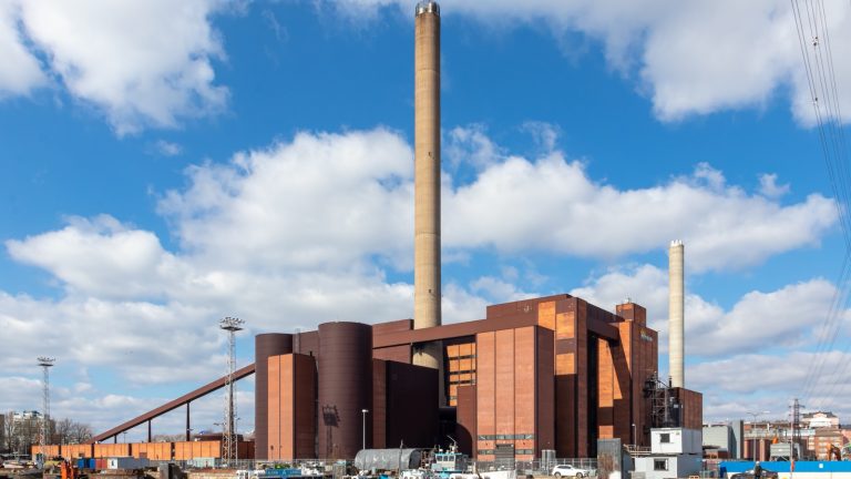 Grande centrale thermique en briques brunes avec une haute cheminée, au bord d'un bassin portuaire où sont amarrés des bateaux, sous un ciel bleu nuageux