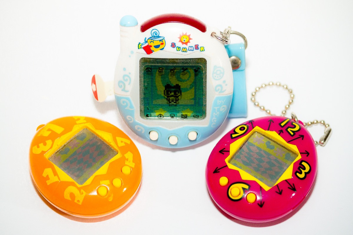 Trois Tamagotchi de modèles différents posés sur un fond blanc : un orange et un rose décoré de chiffres au premier plan, et derrière eux un modèle blanc et bleu en forme de téléphone à antenne, décoré du mot Summer