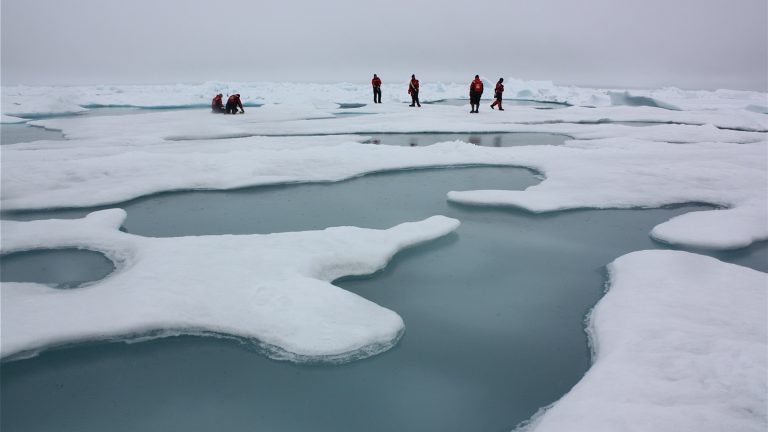 Vaste étendue de glace de mer parcourue de mares de fonte turquoise, avec quelques scientifiques en combinaison rouge au loin