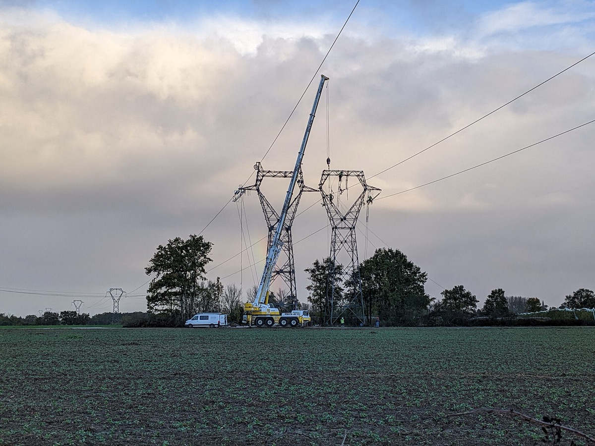 Grue montée à côté de deux pylônes à haute tension au milieu d'un champ, au lever du jour