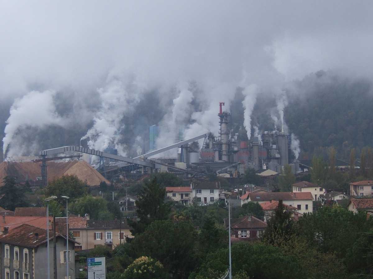 Panaches de vapeur blanche s'élevant au-dessus des bâtiments d'une usine implantée au milieu d'un bourg, par temps couvert