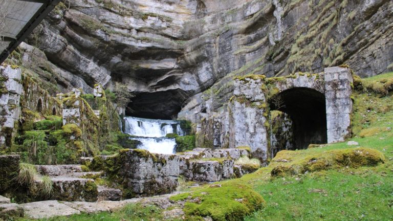Cascade blanche jaillissant d'une grotte au pied d'une haute falaise calcaire couverte de mousse, avec un ancien bâtiment de pierre sur la droite