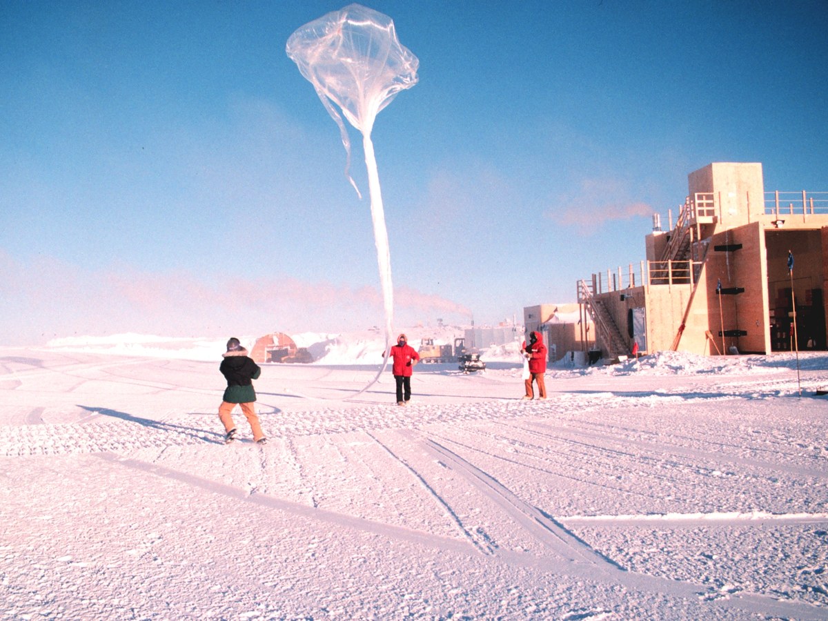 Trois personnes en tenue polaire lâchent un grand ballon transparent sur la neige, devant un bâtiment de station scientifique