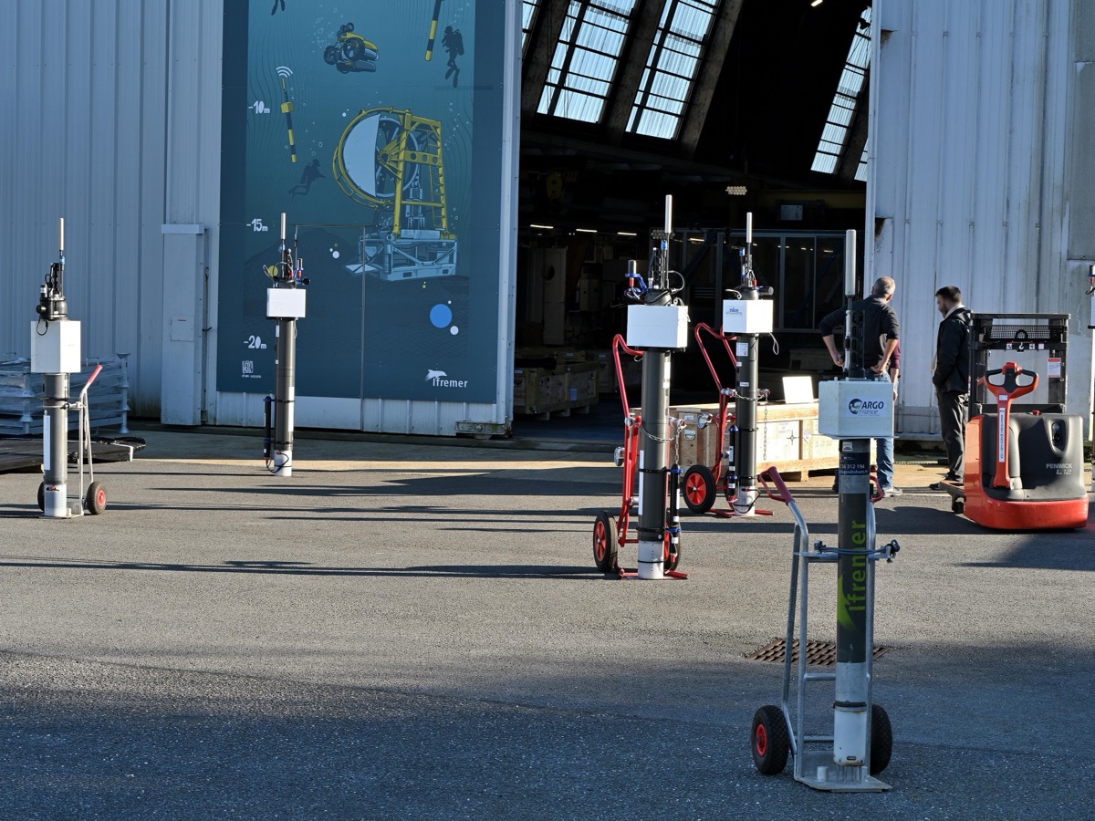 Flotteurs profileurs cylindriques alignés sur un parvis de hangar portuaire, devant des techniciens au travail