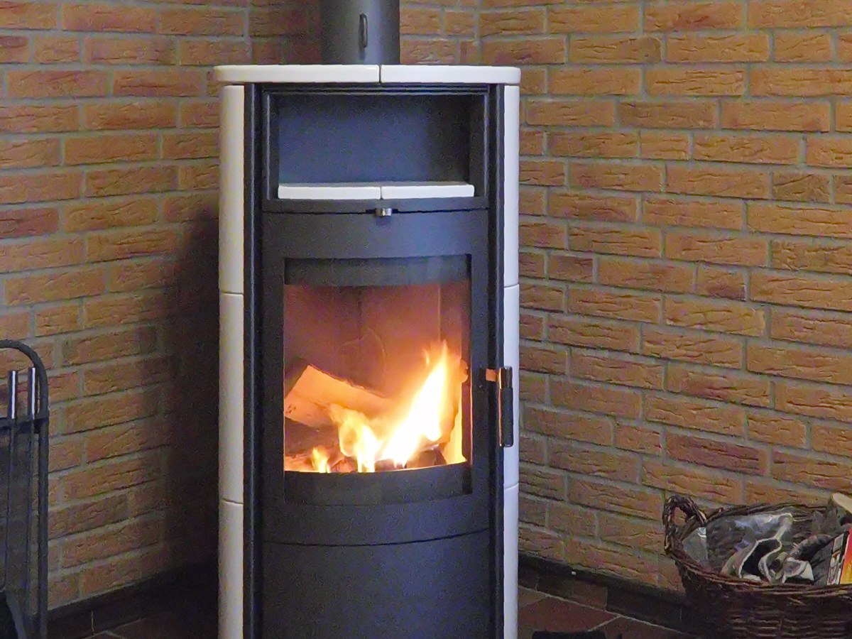 Poêle à bois moderne en fonctionnement dans l'angle d'une pièce aux murs de briques, flammes visibles derrière la vitre, tuyau de raccordement au-dessus