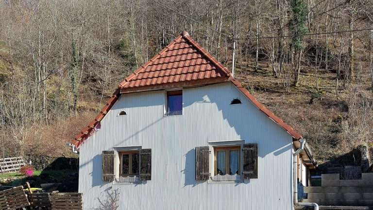 Maison de village aux volets de bois ouverts contre la façade, devant un versant d'arbres sans feuilles en hiver