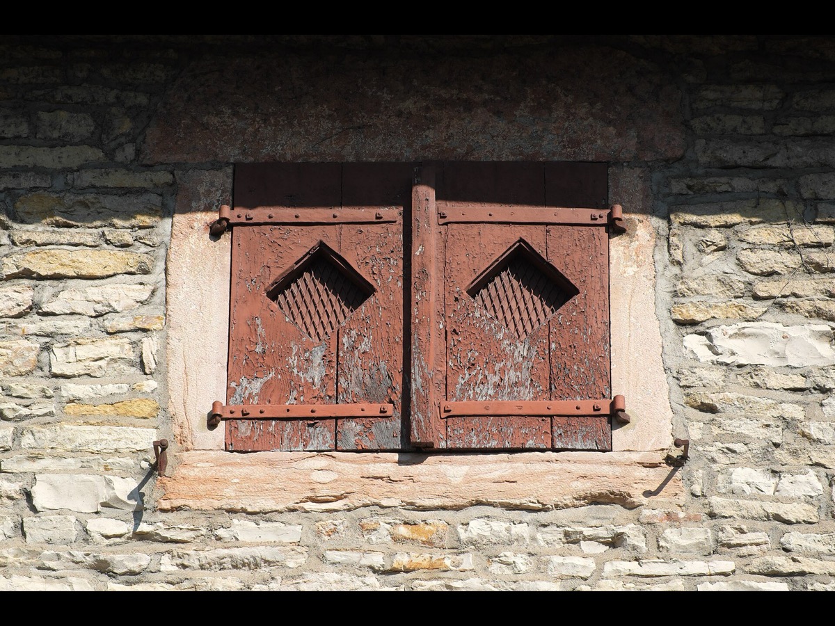 Volets de bois à deux battants fermés sur une fenêtre, peinture écaillée et pentures métalliques, dans un mur de pierre