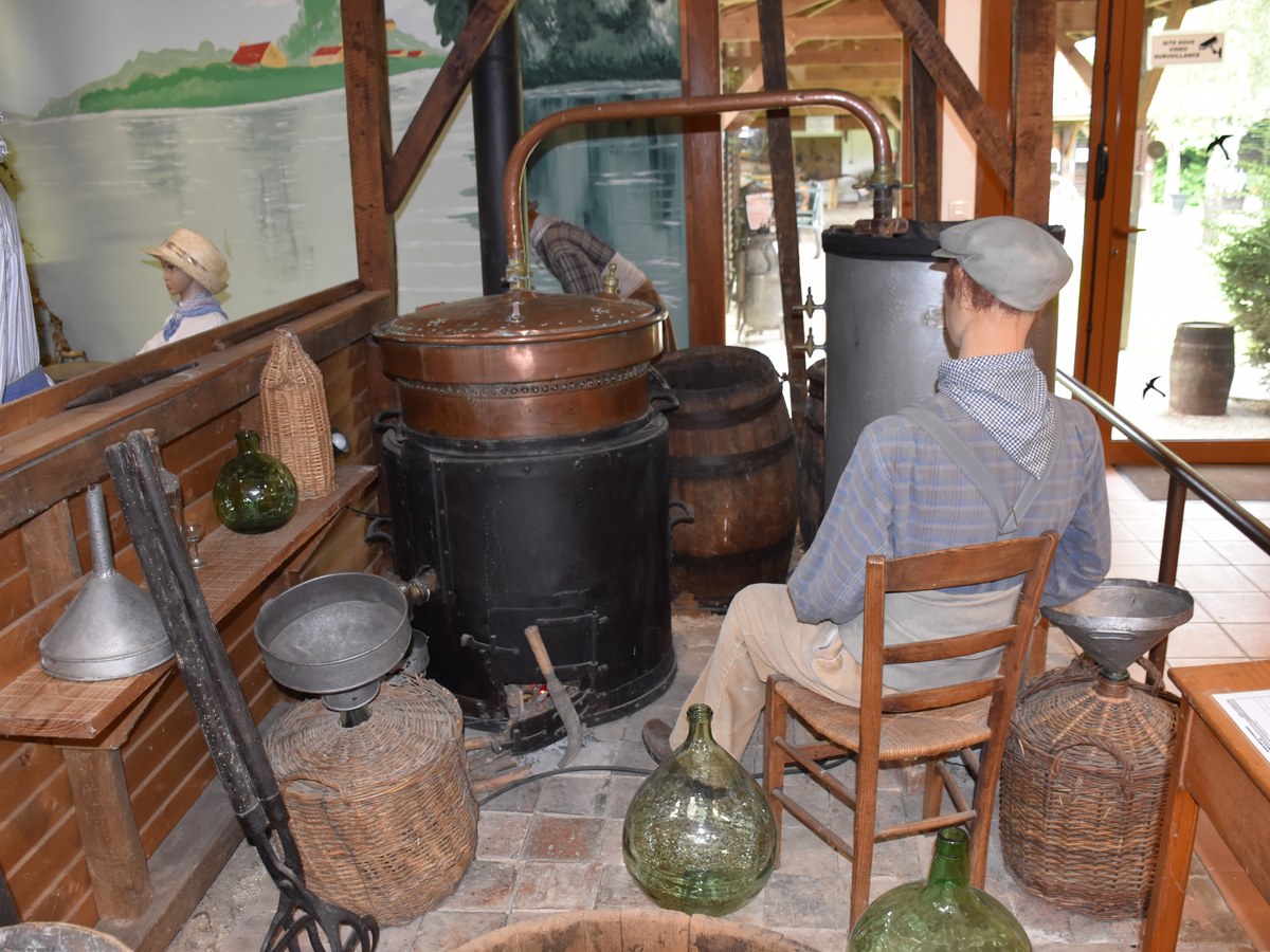 Reconstitution d'un atelier de distillation avec un alambic de cuivre, un mannequin en casquette assis devant, des dames-jeannes en verre et des entonnoirs de métal