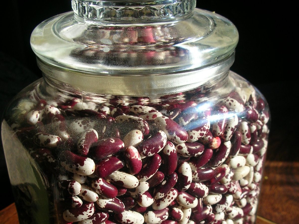 Grand bocal en verre à couvercle rodé rempli de haricots secs pourpres et blancs, posé sur un meuble en bois