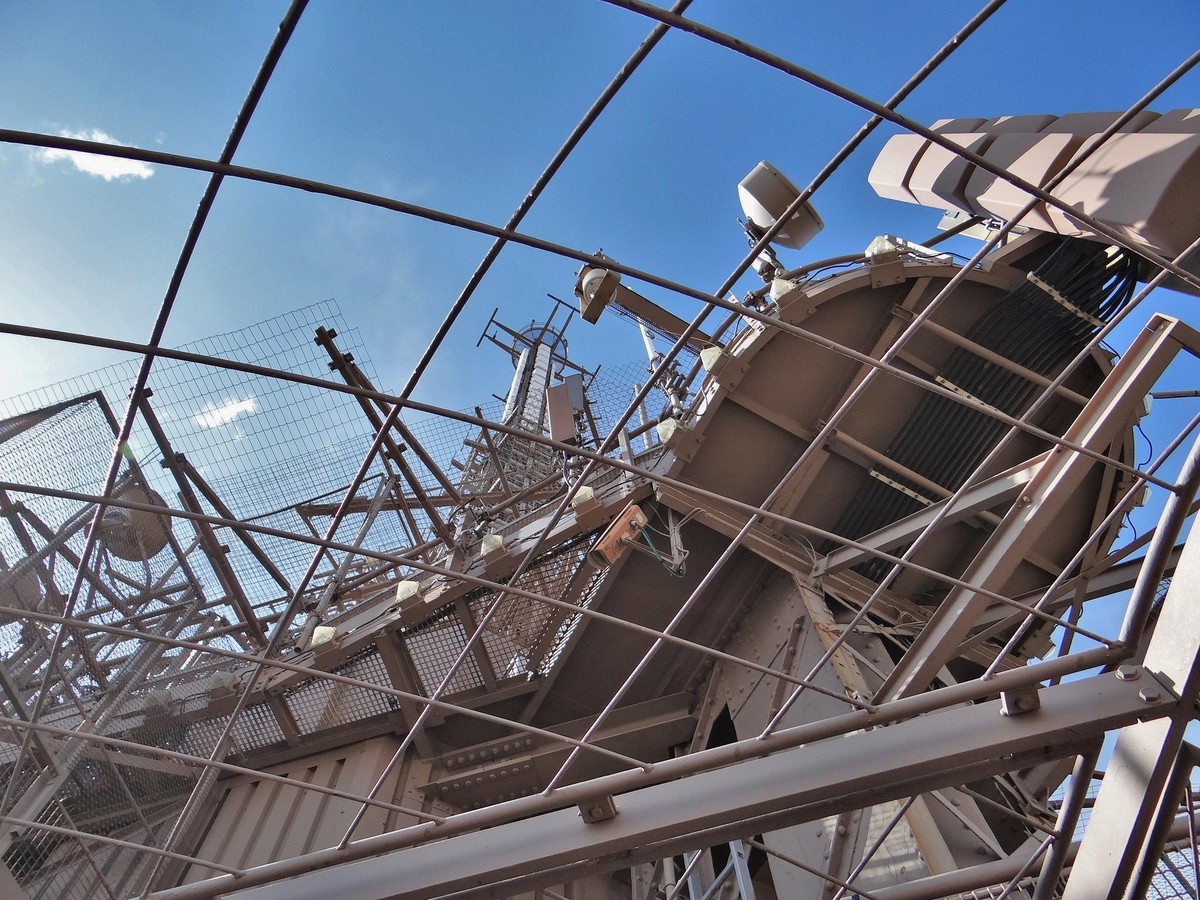 Structure métallique et antennes au sommet de la tour Eiffel vues en contre-plongée sous un ciel bleu, derrière un grillage de protection