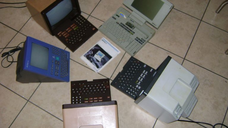 Six terminaux Minitel de modèles et de couleurs différents posés au sol sur un carrelage clair, écrans éteints et claviers dépliés