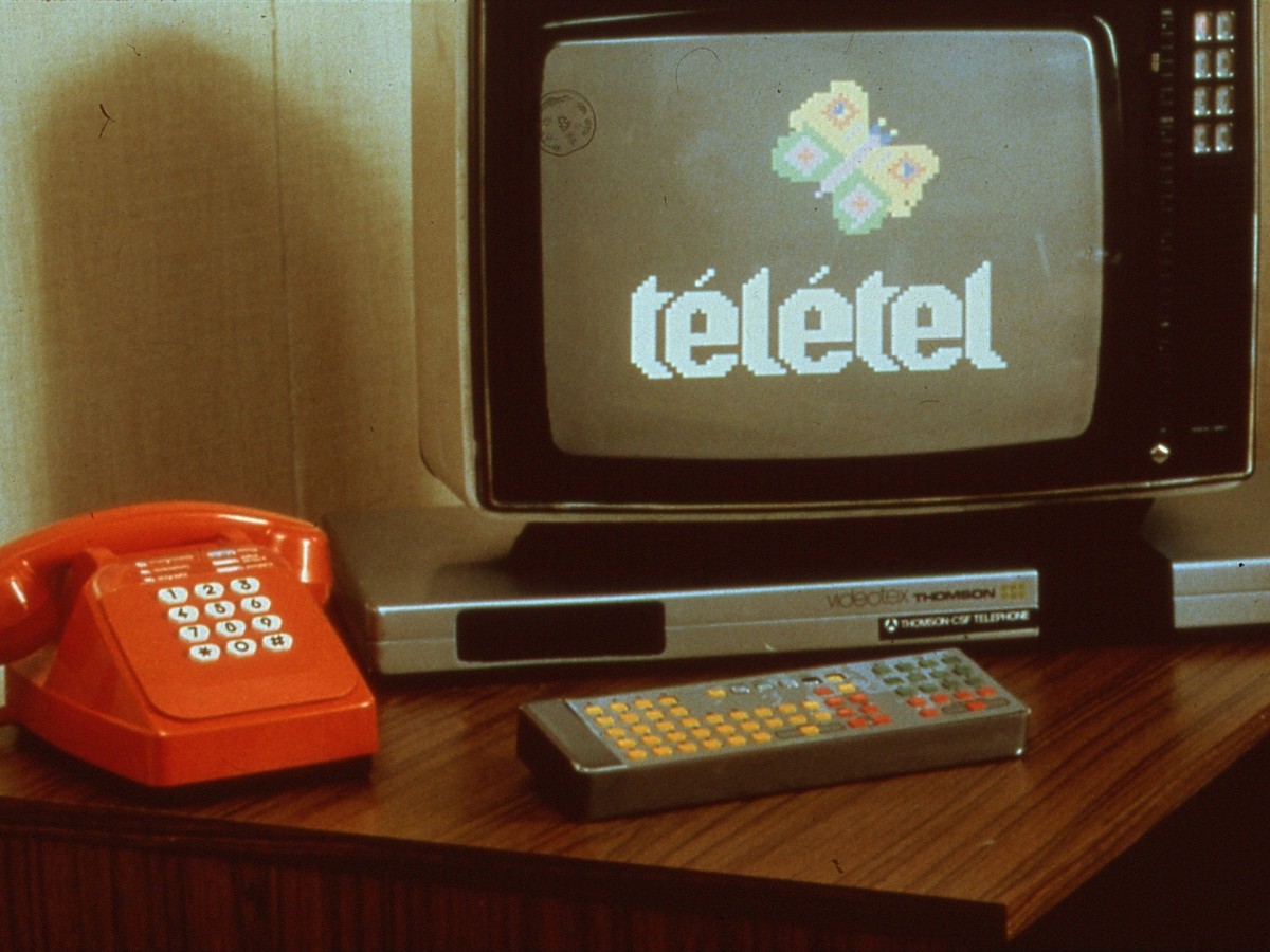 Terminal de vidéotex Thomson posé sur un meuble en bois, un téléphone à touches rouge à sa gauche et un clavier plat argenté posé devant lui