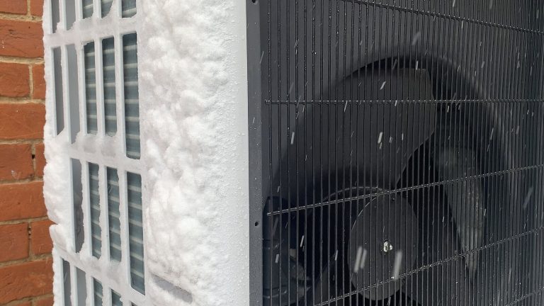 Unité extérieure de pompe à chaleur fixée contre un mur de brique, sa grille latérale couverte de neige, pendant une chute de neige
