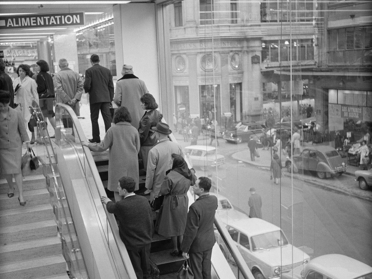 Escalier mécanique d’un magasin parisien en 1965, clients en manteau montant vers un étage signalé Alimentation, avec la rue et les Magasins du Printemps visibles derrière la vitre