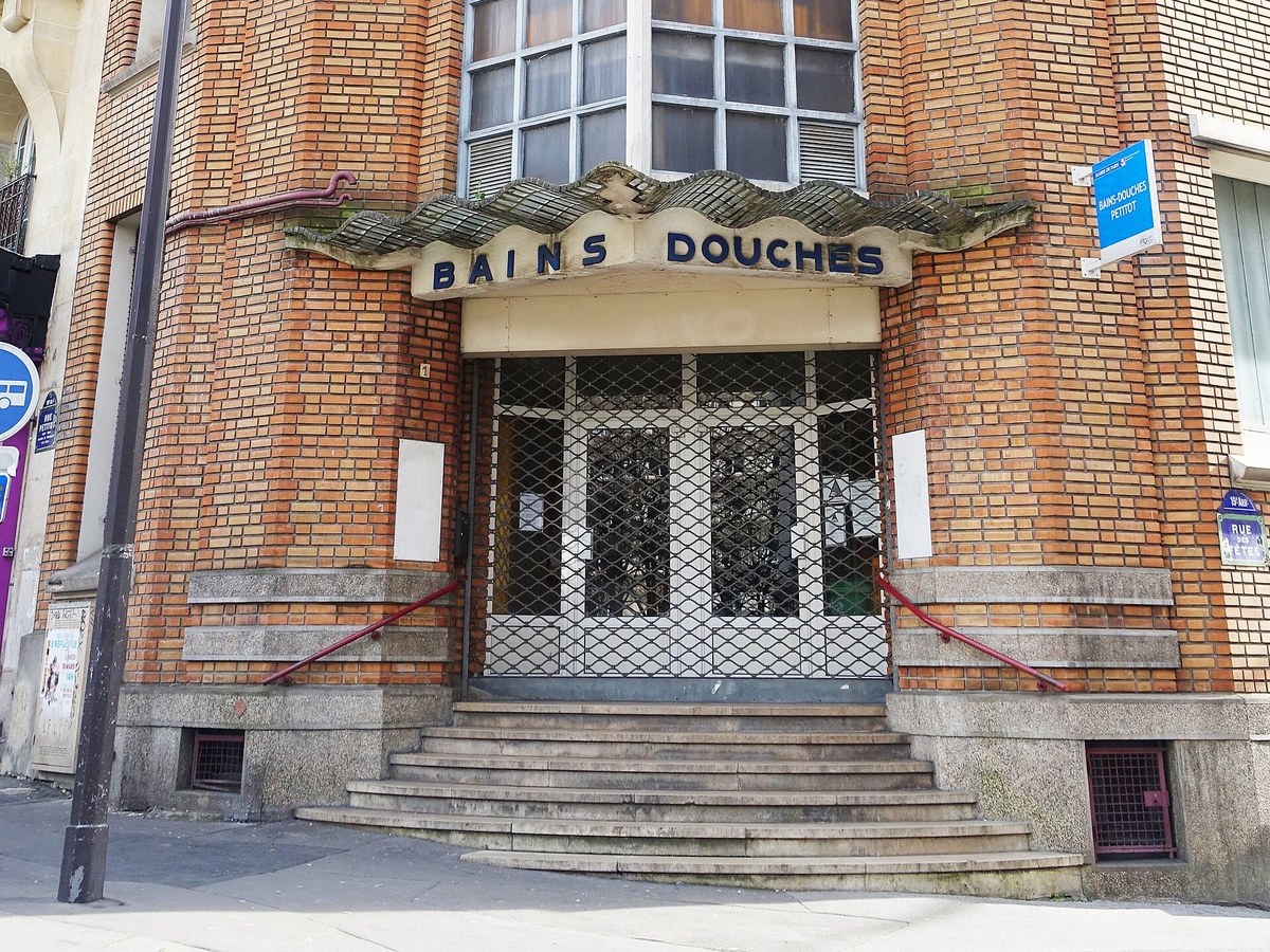 Entrée en brique d'un bâtiment des années 1930 surmontée d'une marquise ondulée portant les lettres bleues Bains Douches, avec un escalier de pierre et une grille métallique