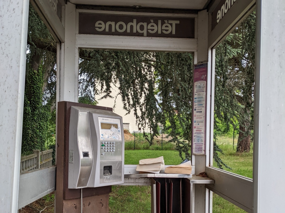 Intérieur d’une cabine téléphonique vitrée avec un publiphone à carte argenté sans combiné, des annuaires abîmés posés sur la tablette et de la végétation derrière les vitres